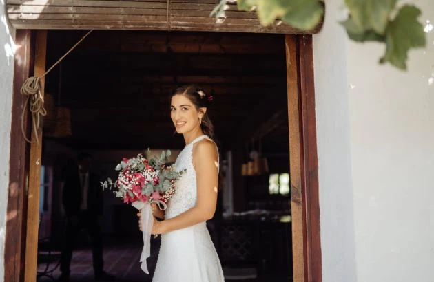 Novia con un vestido de encaje y ramo de flores de pie bajo una pérgola rústica en un soleado día de boda.